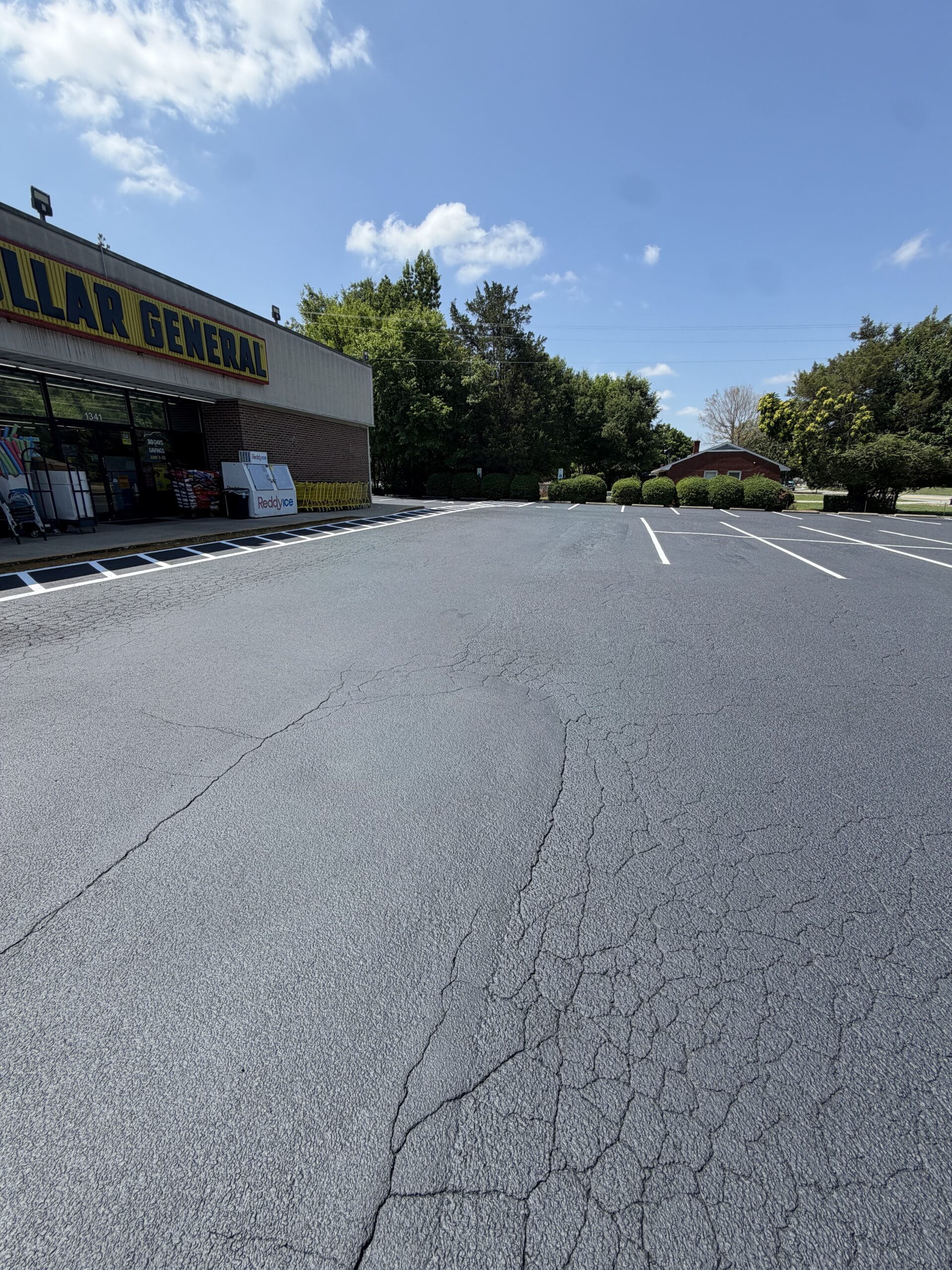 Front view of a Dollar General store with a mostly empty parking lot under a blue sky and trees behind.