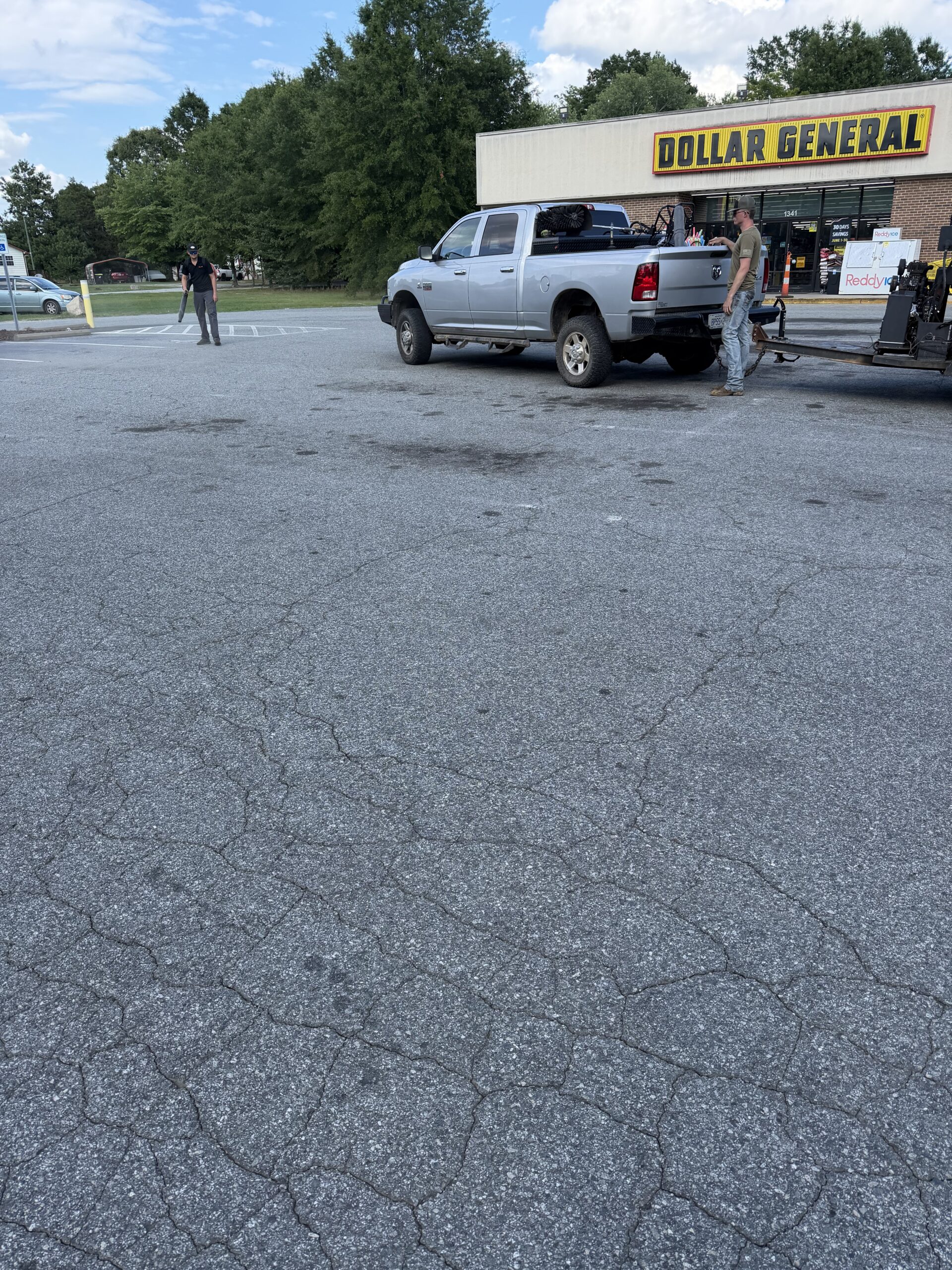 Silver pickup with trailer attached in a parking lot in front of a Dollar General store; two men nearby inspecting equipment on the trailer.