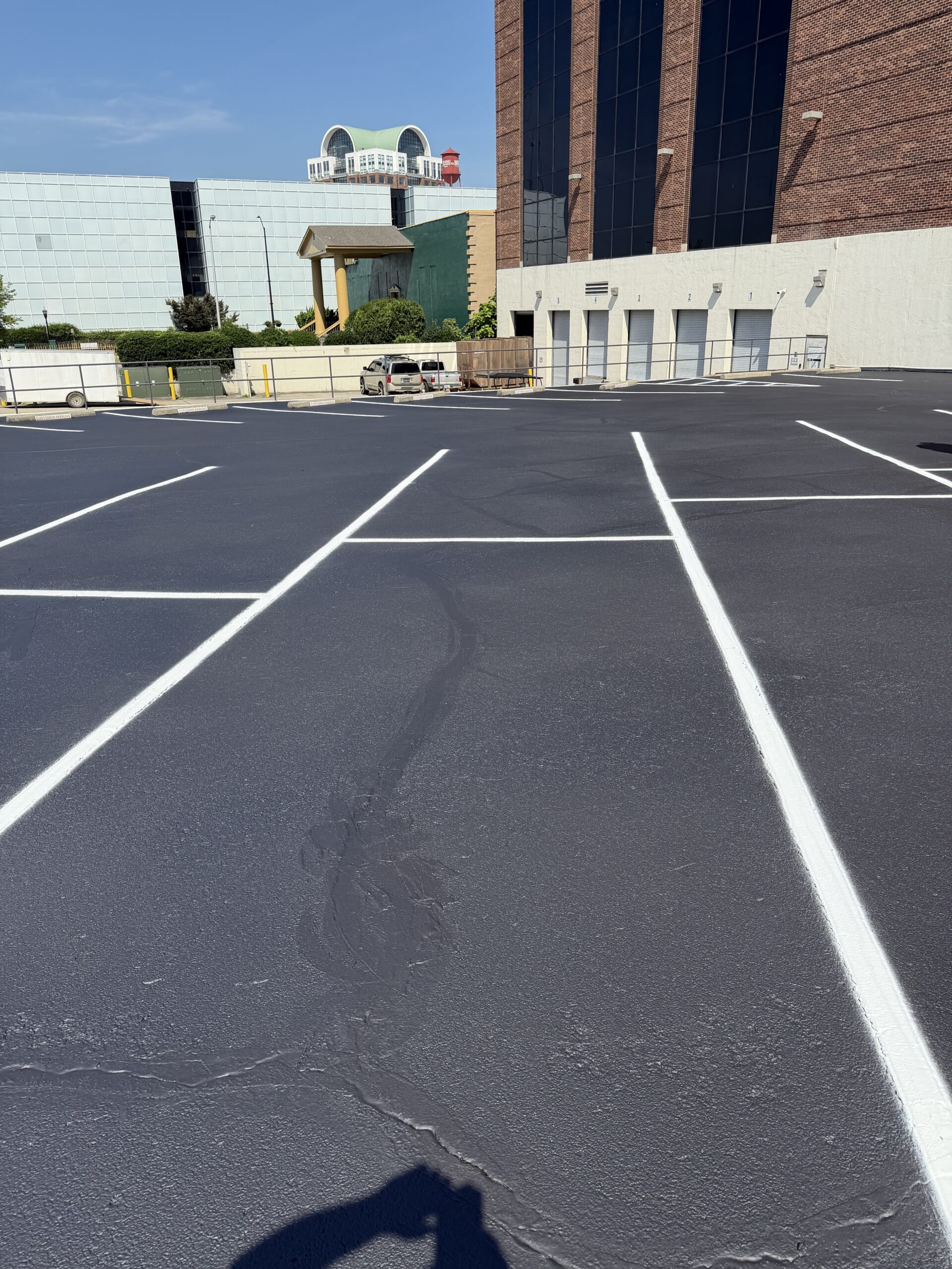 Wide view of a paved parking lot with white lines, a brick building on the right, a beige wall with garage doors, and a few parked cars under a clear blue sky.