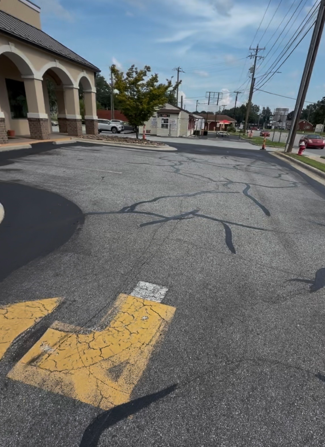 Parking lot outside a shopping center with cracked pavement, patches, and faded yellow turn-arrow markings near a building with arched columns on the left.